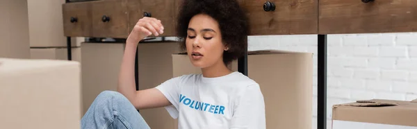 Tired african american woman sitting near rack with boxes in volunteer center, banner — Stockfoto