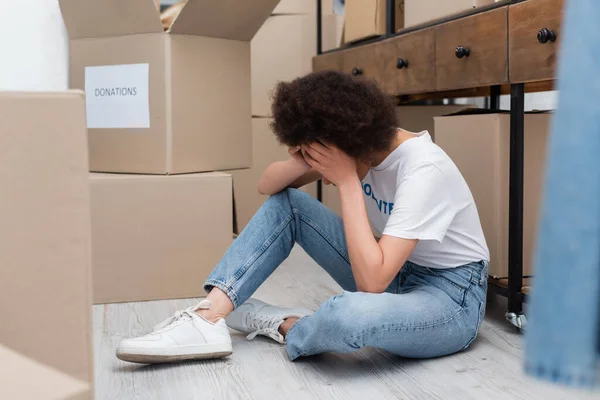 Exhausted african american woman obscuring face with hands while sitting on floor in volunteer center — Photo de stock