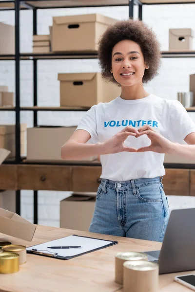 Feliz africano americano voluntario mostrando corazón signo cerca portapapeles en escritorio - foto de stock