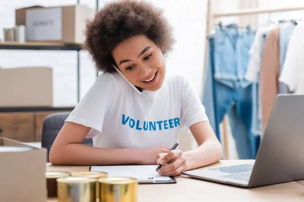 Sorrindo africano americano voluntário falando no smartphone e escrevendo perto de laptop no centro de caridade — Fotografia de Stock