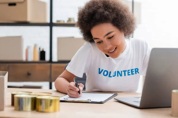 Smiling african american volunteer writing on clipboard near blurred laptop — Stock Photo