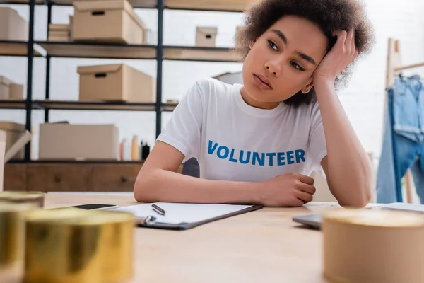 Cansado y pensativo afroamericano voluntario sentado en el lugar de trabajo en el centro de caridad - foto de stock