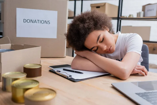 Exhausted african american volunteer sleeping near smartphone and blurred canned food - foto de stock