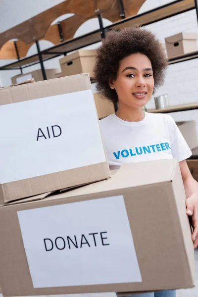 Africano americano voluntário olhando para câmera perto de caixas com ajuda e doar letras — Fotografia de Stock