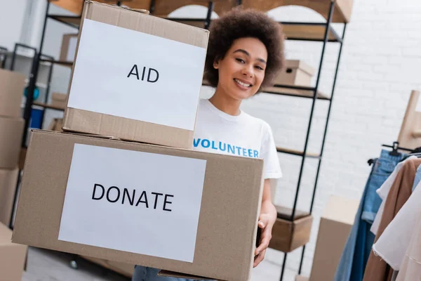 Africano americano voluntário sorrindo perto de caixas de papelão com doar e ajuda lettering — Fotografia de Stock