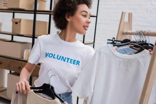 Smiling african american volunteer holding hangers with clothes in charity storehouse — Stock Photo