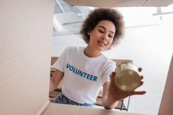 Bottom view of smiling african american woman with tin near carton box — Foto stock