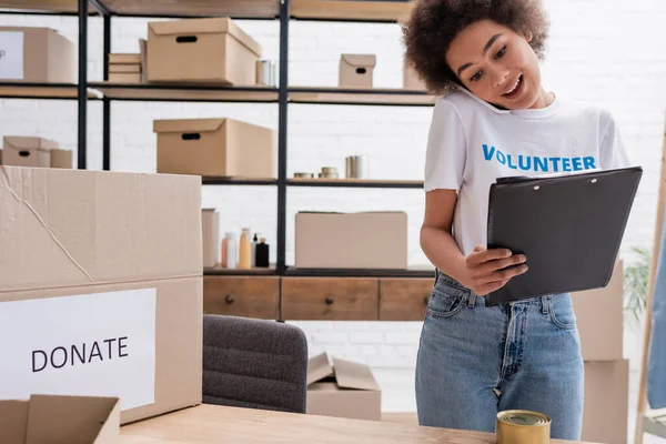 African american volunteer talking on mobile phone near clipboard in charity warehouse — Stock Photo