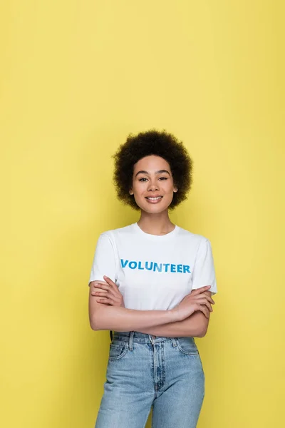 Pleased african american woman with volunteer lettering on t-shirt standing with crossed arms isolated on yellow — Stock Photo