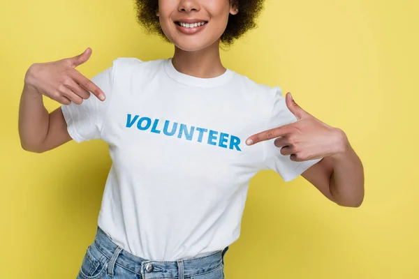 Vue recadrée d'une femme afro-américaine souriante pointant du doigt une inscription bénévole sur un t-shirt isolé sur du jaune — Photo de stock