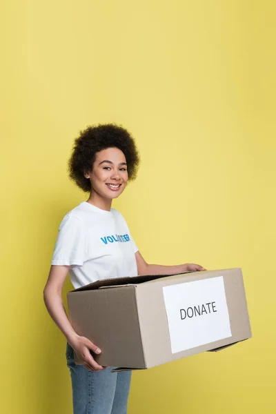 Happy african american woman with donate box looking at camera isolated on yellow — Stock Photo