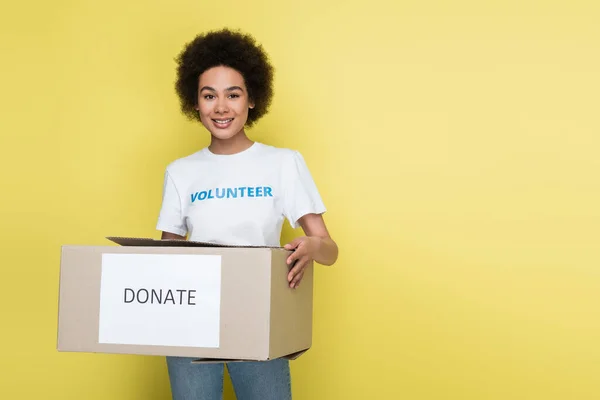 Позитивный африканский доброволец держит коробку пожертвований изолированной на желтый — стоковое фото