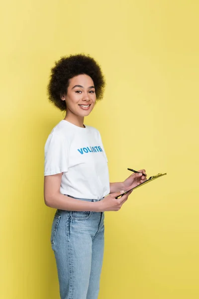 Smiling african american volunteer writing on clipboard isolated on yellow — Stockfoto