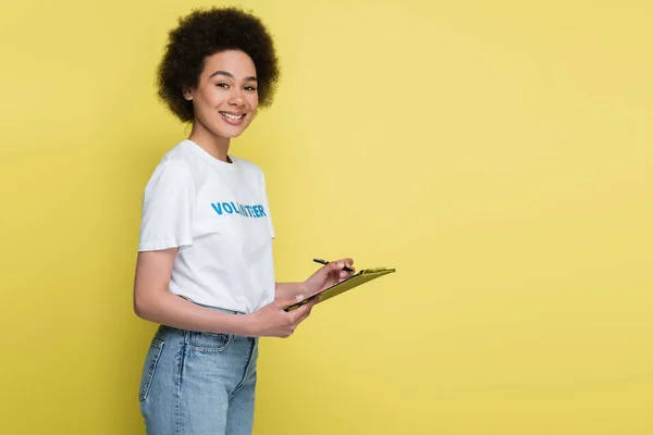 Young african american volunteer writing on clipboard and looking at camera isolated on yellow — Stockfoto