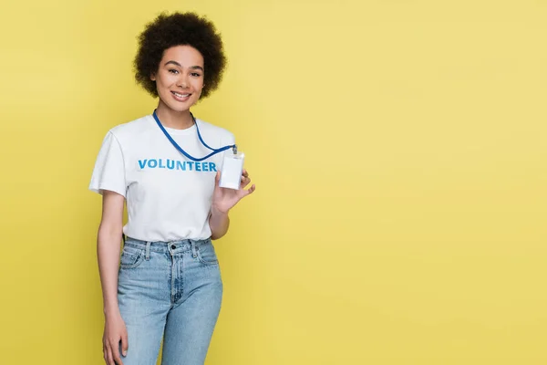 Voluntario afroamericano sonriendo a la cámara y mostrando tarjeta de identificación en blanco aislado en amarillo - foto de stock