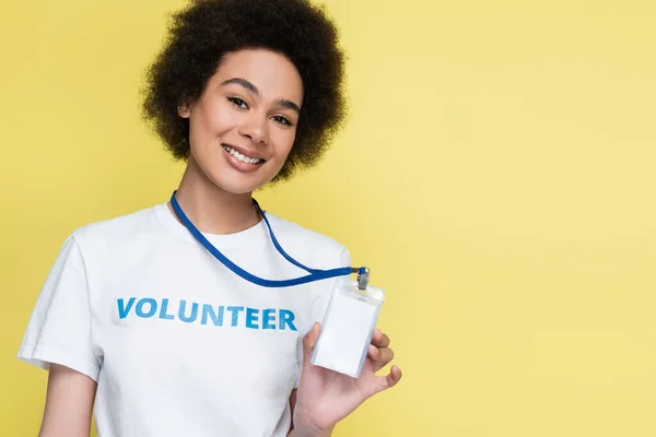 Voluntario afroamericano sonriendo a la cámara y mostrando tarjeta de identificación en blanco aislado en amarillo - foto de stock