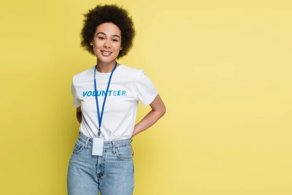 Sonriente afroamericano voluntario con etiqueta de nombre en blanco y las manos detrás de la espalda aislado en amarillo - foto de stock