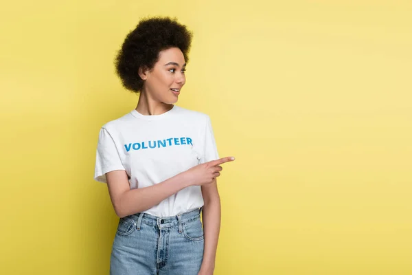 African american woman in t-shirt with volunteer lettering pointing with finger isolated on yellow — Stockfoto