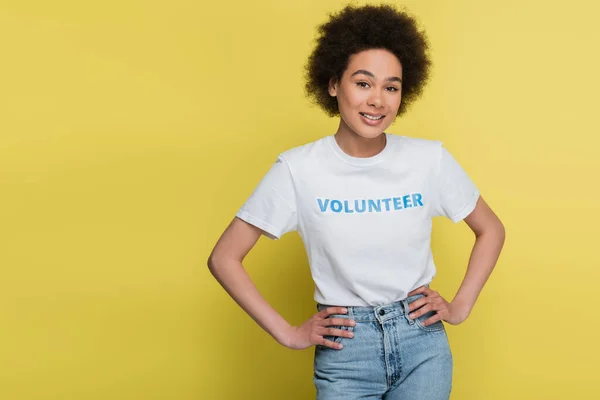 Happy african american woman in t-shirt with volunteer lettering standing with hands on hips isolated on yellow — Stock Photo