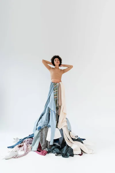 Trendy african american woman posing on pile of clothes with hands behind head on grey background — Foto stock