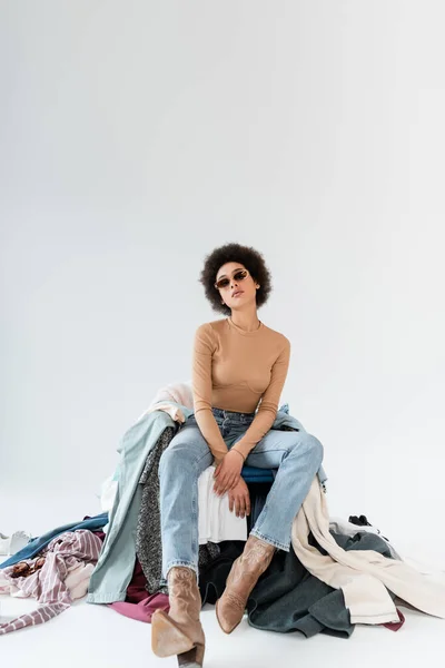 Young african american woman sitting in sunglasses and jeans near mess of clothes on grey background — Stockfoto