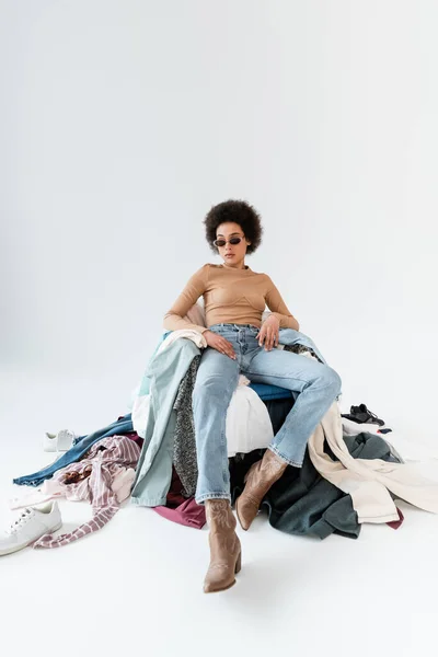 Afro-américaine dans des lunettes de soleil à la mode assis près de différents vêtements sur fond gris — Photo de stock