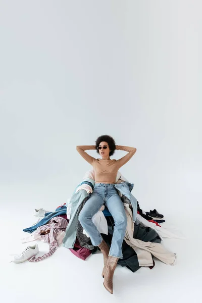 African american woman with hands behind head sitting near mess of clothes on grey background - foto de stock