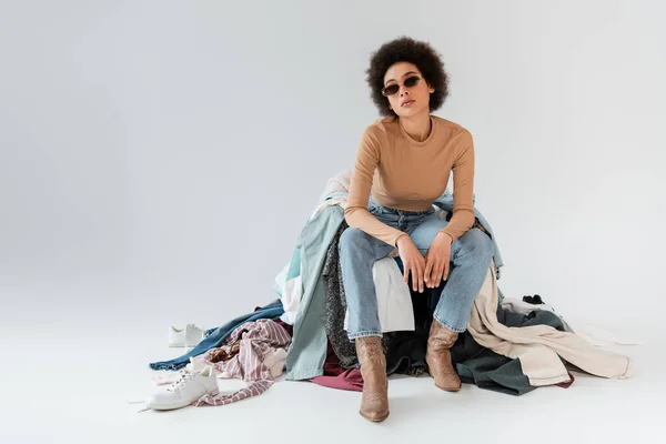 Mulher americana africana na moda em óculos de sol sentado perto de bagunça de roupas diferentes no fundo cinza — Fotografia de Stock