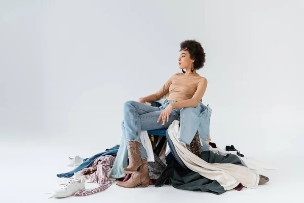 Stylish african american woman in jeans sitting near pile of clothing on grey background — Stock Photo