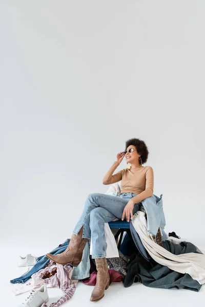 Happy african american woman in jeans and sunglasses sitting near plenty of clothes  on grey background — Stock Photo