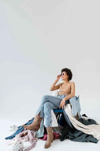 Mujer afroamericana feliz en jeans y gafas de sol sentado cerca de un montón de ropa sobre fondo gris - foto de stock