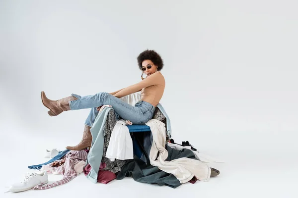 Elegante donna afroamericana in occhiali da sole e stivali cowboy in posa vicino mucchio di abbigliamento su sfondo grigio — Foto stock