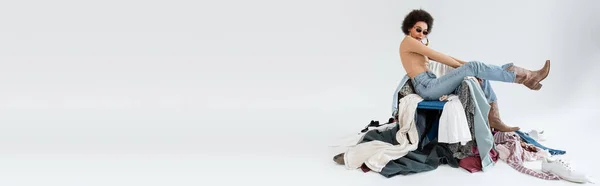 Full length of african american woman sitting near pile of clothing on grey background, banner — Stock Photo