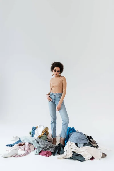 Pleine longueur de femme afro-américaine posant avec le pouce dans la poche de jeans près pile de vêtements sur fond gris — Photo de stock