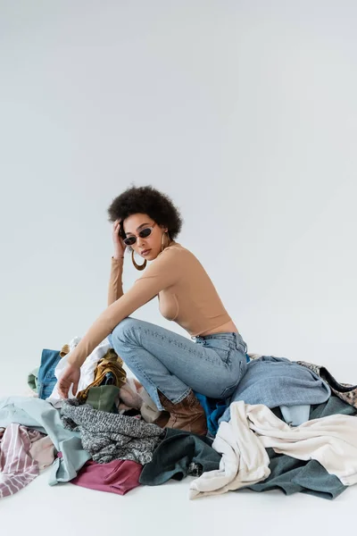 Curly african american woman in sunglasses posing near assortment of clothes on grey background — Stock Photo