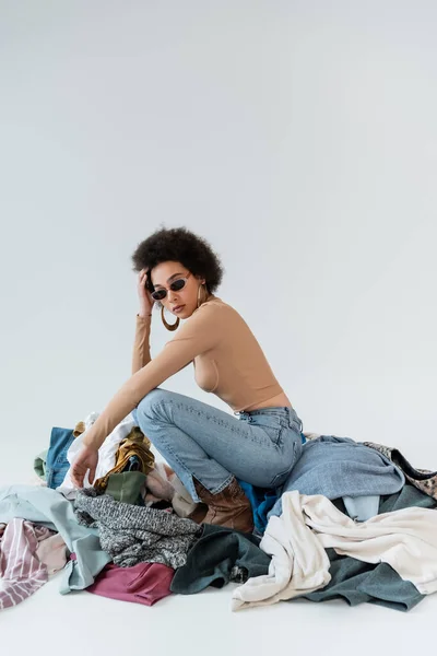 Mujer americana africana rizada en gafas de sol posando cerca de surtido de ropa sobre fondo gris - foto de stock