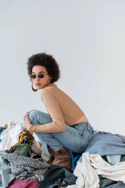 Brunette african american woman in trendy sunglasses near pile of different clothes on grey background — Stockfoto