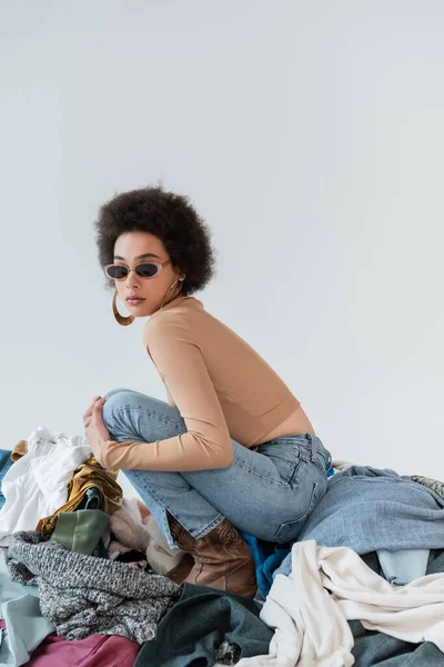 Brunette african american woman in trendy sunglasses near pile of different clothes on grey background — Stock Photo