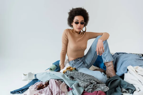 African american woman in stylish sunglasses sitting on variety of clothes on grey background - foto de stock