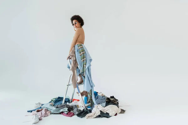 Fashionable african american woman standing on ladder above mess of garments on grey background — Stockfoto