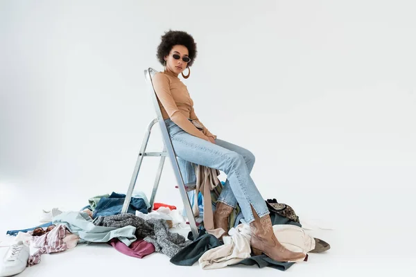 African american woman sitting on ladder near mess of garments on grey background — Fotografia de Stock
