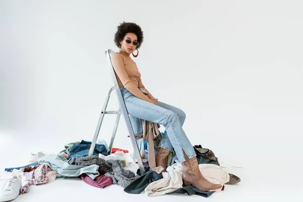 Afro-américaine femme assise sur l'échelle près mess de vêtements sur fond gris — Photo de stock