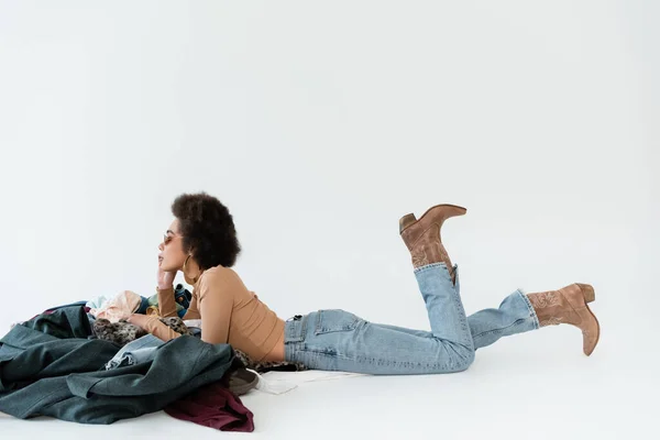 Vista lateral de la mujer afroamericana en jeans y botas de vaquero acostado cerca de un montón de ropa sobre fondo gris - foto de stock