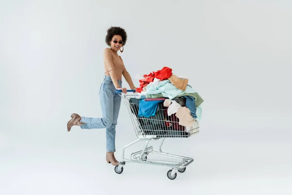 Full length of happy african american woman near shopping cart full of clothes on grey background — Fotografia de Stock