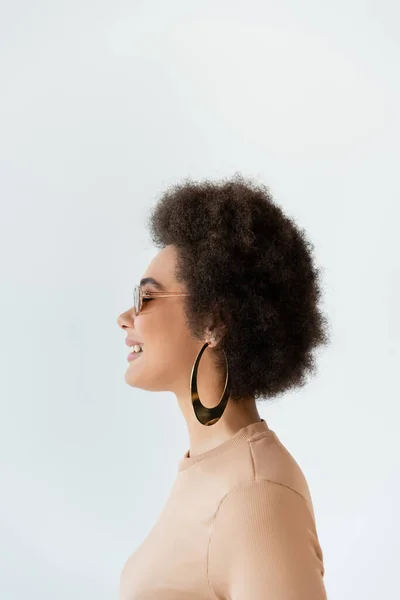 Вид збоку кучерявої афроамериканської жінки в сережці та сонцезахисних окулярах, посміхаючись ізольовано на сірому — стокове фото