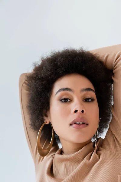 Retrato de mujer afroamericana en pendientes de anillo posando con las manos detrás de la cabeza aisladas en gris - foto de stock