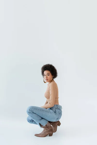 Trendy african american woman in beige long sleeve shirt and jeans posing on grey background — Stockfoto