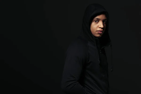 Sudadera afroamericana con capucha mirando a la cámara mientras está de pie aislado en negro - foto de stock