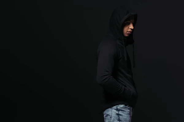 Side view of african american hooligan in hoodie standing isolated on black — Fotografia de Stock