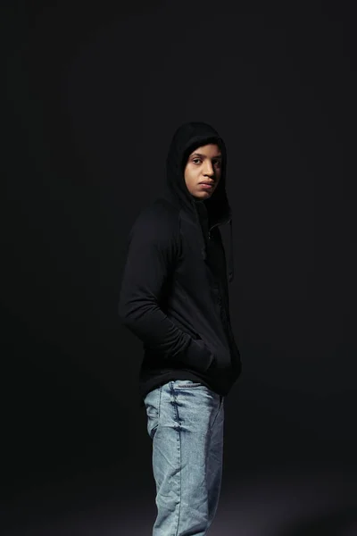 African american hooligan holding hand in pocket of hoodie isolated on black — Photo de stock